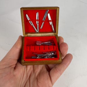 May include: A miniature wooden box with a red velvet interior, holding tiny silver cutlery. The box contains miniature knives, forks, and spoons, perfect for a dollhouse or miniature collection. The box is held in a hand.