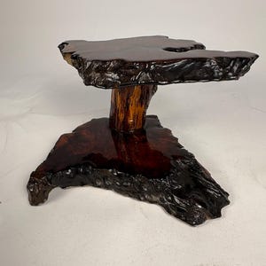 May include: A handcrafted wooden display stand with a dark, glossy finish. The stand features a natural, irregular shape with a dark brown top and base, supported by a lighter brown, textured trunk-like pillar. The wood grain is visible, adding to its rustic charm.