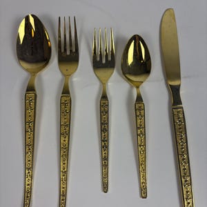 May include: A set of five gold-colored flatware pieces: a spoon, two forks, another spoon, and a knife. The handles are decorated with a repeating floral pattern. The flatware is arranged on a white surface.
