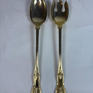 May include: A gold-colored serving spoon and fork set. The spoon has a rounded bowl, and the fork has three tines. Both utensils have long, slender handles with ornate, shell-like designs at the base.