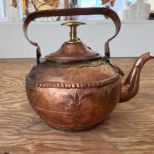 May include: A copper teapot with a curved handle and spout. The teapot has an ornate, etched floral design around the body and a brass-colored top and handle base. The teapot sits on a wooden surface.