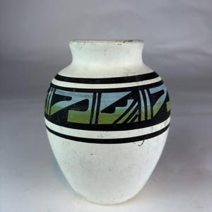 May include: A white ceramic vase with a rounded body and a narrow neck. The exterior features a horizontal band decorated with black geometric patterns and a gradient of blue and green colors painted within the shapes around the center.