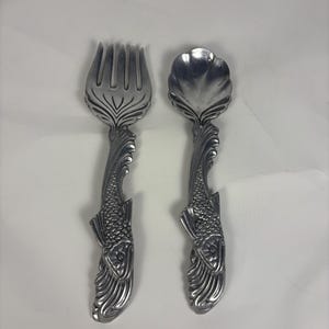 May include: A silver-colored fork and spoon set. The handles are designed with a fish motif, featuring detailed scales and fins. The fork has four tines, and the spoon has a shell-shaped bowl. The set is likely for serving or decorative use.