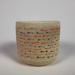 May include: A beige ceramic cup with a rounded base. The cup is decorated with a pattern of small, horizontal orange and gray dashes. The cup has a textured surface and a simple, minimalist design.