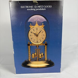 May include: A gold-toned electronic quartz clock with a revolving pendulum, encased in a glass dome. The clock face has black Roman numerals and gold hands. The box reads "ELECTRONIC QUARTZ CLOCKS revolving pendulum".