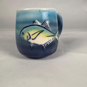May include: Ceramic mug with a hand-painted fish design. The mug features a gradient of blue hues, transitioning from light blue at the top to a deep navy at the base. The fish is depicted in shades of yellow, white, and black.
