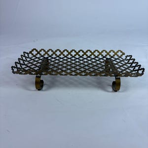 May include: A vintage brass-toned metal shelf with a decorative openwork design. The shelf has a rectangular shape with a repeating quatrefoil pattern and curved supports. It is likely used for displaying small items.