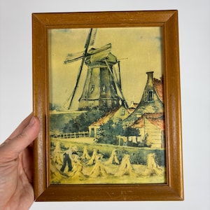 May include: A framed print of a rural landscape featuring a windmill, houses, and a field of haystacks. The artwork is set in a wooden frame. The color palette includes shades of yellow, green, and brown.