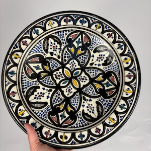 May include: A decorative ceramic plate with a detailed, symmetrical design. The plate features a black rim and a central pattern in blue, white, yellow, and red. The intricate design is reminiscent of traditional pottery.
