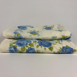 May include: Two folded cream-colored bed sheets with a repeating pattern of blue roses and green leaves. The sheets are stacked on top of each other, showcasing the floral design.