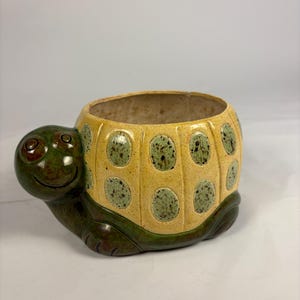 May include: A ceramic turtle-shaped planter. The turtle has a yellow shell with green polka dots, a green head, and green feet. The planter has a smiling face and a large opening for plants. The planter is approximately 6 inches long.