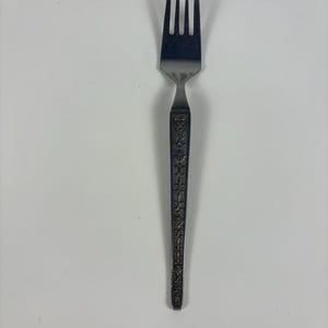 May include: A stainless steel fork with four tines. The handle features an ornate floral design. The fork is designed for eating and is a piece of flatware.