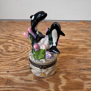 Puede incluir: Caja de baratijas decorativa con dos orcas en blanco y negro sobre un arreglo floral. La caja es redonda, con tapa con bisagras y base texturizada. Hecha de cer&aacute;mica.