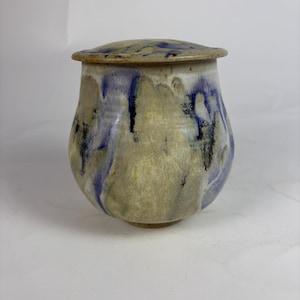 May include: A handmade ceramic jar with a lid. The jar is a light beige color with blue and black streaks. The lid is a similar color with blue and black accents. The jar has a rounded shape.
