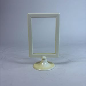 May include: A cream-colored rectangular picture frame with a rounded base. The frame is empty, and the stand is a matching cream color. The frame is approximately 6 inches tall and 4 inches wide.
