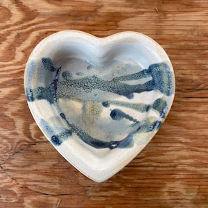 May include: Heart-shaped ceramic dish with a white base and blue and teal abstract design. The interior features a textured, speckled pattern. The dish is placed on a wooden surface.