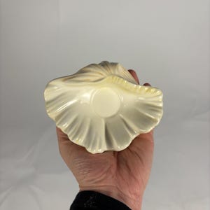May include: A cream-colored, seashell-shaped ceramic dish. The dish has a ruffled edge and a small, circular indentation in the center. The exterior has a textured, ridged design. The dish is held in a hand against a plain background.
