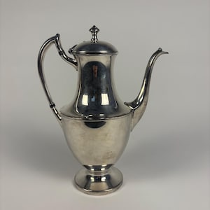 May include: A silver-colored coffee pot with a curved spout and handle. The pot has a lid with a small decorative finial. The base is a pedestal design. The pot is likely made of metal and reflects the surrounding environment.