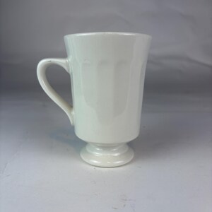 May include: A white ceramic mug with a glossy finish, featuring a vertical fluted pattern around the upper body, a curved handle on the left side, and a pedestal base that elevates the main cup section from the surface it rests upon.