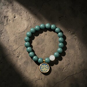 Pode incluir: Uma pulseira de contas turquesa com um pingente dourado com um design azul e amarelo. A pulseira inclui uma única conta branca. A joia é exibida em uma superfície marrom texturizada.
