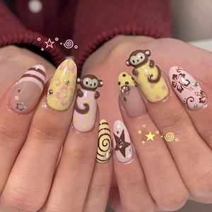 May include: Oval nail art with diverse designs. Some nails feature pink and brown stripes, yellow with pink floral patterns, and monkey figures. Accents include stars and rhinestones, creating a playful aesthetic.