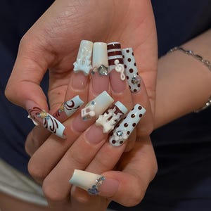 Puede incluir: Uñas postizas largas en forma de ataúd con una mezcla de diseños que incluyen degradados en azul claro y blanco, lunares marrones, rayas horizontales marrones y dijes 3D blancos de notas musicales, acentuados con pequeños brillantes.