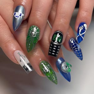 May include: Stiletto-shaped artificial nails featuring diverse designs including a metallic blue cat face, green circuit board patterns, a black vintage mobile phone, a blue globe, white wired earbuds, and a white nail with a silver sword icon.