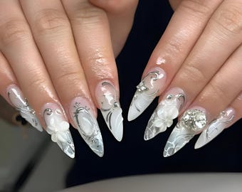 May include: Long, pointed acrylic nails with intricate designs. The nails feature a white base with silver embellishments, including floral accents and decorative swirls. Some nails have a clear, marbled effect.