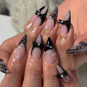 May include: Long, pointed acrylic nails with a black and lace design. The nails feature black tips, lace accents, small black bows, and pearl embellishments. The base of the nails is a natural pink color.