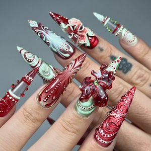 Op de afbeelding: Een set lange, puntige acrylnagels met ingewikkelde ontwerpen. De nagels hebben een combinatie van rode, witte en groene kleuren, met bloemen- en abstracte patronen. Sommige nagels hebben 3D-versieringen.