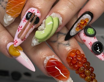 May include: Stiletto-shaped nails featuring 3D sushi-themed art. Designs include orange salmon slices, green avocado, glossy red fish roe, black seaweed wraps, and brown chopsticks on a light pink base with small red floral patterns.