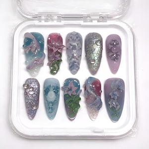 May include: A collection of ten artificial nails presented in a clear case. Each nail showcases a distinct design, incorporating shades of blue, purple, and green, with floral and abstract decorations. The nails have a glossy finish.