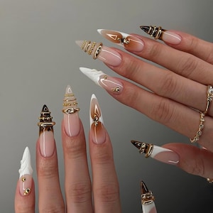 Puede incluir: Uñas acrílicas largas en forma de estilete, con un diseño degradado de nude a blanco. Anillos y tachuelas doradas adornan las uñas, añadiendo un toque de glamour. Las uñas son un accesorio de moda.