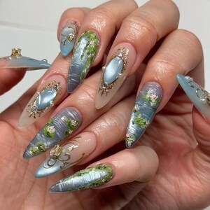 May include: A hand with long, almond-shaped nails featuring intricate nail art. Designs include water lily ponds, metallic accents, and ornate, oval-shaped gems. The color palette is soft, with blues, greens, and gold.