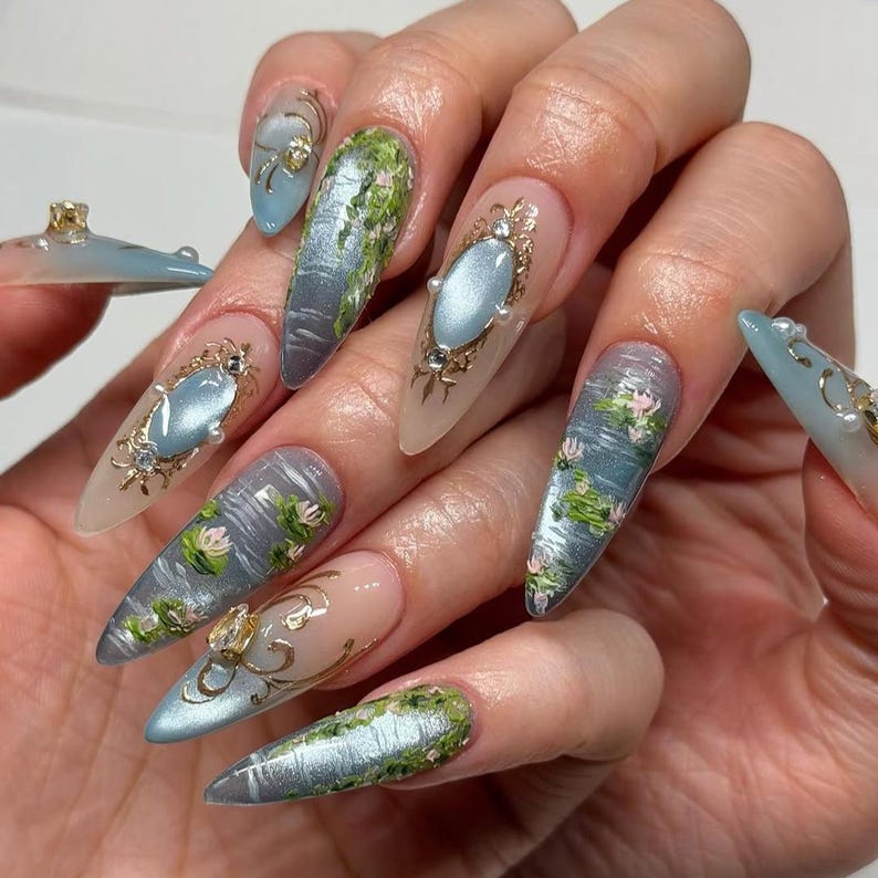 May include: A hand with long, almond-shaped nails featuring intricate nail art. Designs include water lily ponds, metallic accents, and ornate, oval-shaped gems. The color palette is soft, with blues, greens, and gold.