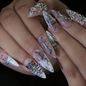 May include: Long, pointed acrylic nails with a white and clear ombre design. The nails feature silver embellishments, including floral accents, studs, and intricate black and silver line art. Some nails have text.