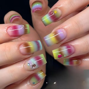May include: Square acrylic nails with a gradient effect in pink, yellow, green, and brown. Some nails are decorated with small gems. The nails have a glossy finish. The nails are well-manicured.