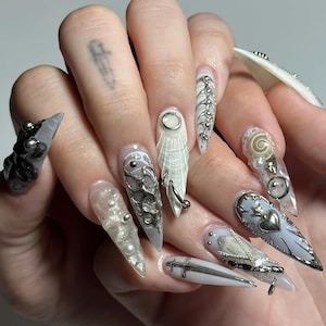 May include: A hand displaying a set of long, pointed, artificial nails. The nails feature a variety of designs, including silver embellishments, pearls, and intricate patterns. Some nails are clear, while others are painted in shades of white and gray.