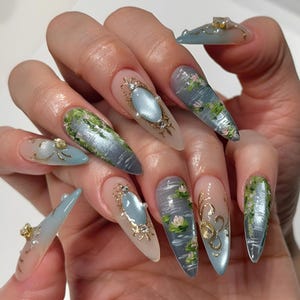 May include: A hand displays long, almond-shaped acrylic nails. Each nail features a unique design, including water scenes with lily pads, ornate gold frames with blue gems, and green mossy textures. Some nails have gold embellishments.