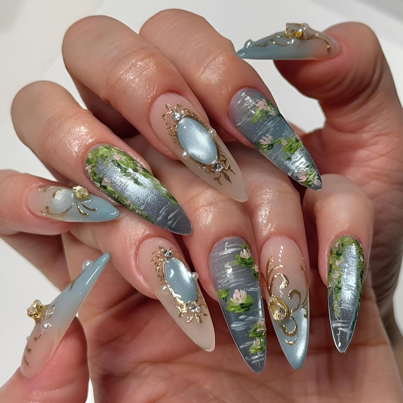 May include: A hand displays long, almond-shaped acrylic nails. Each nail features a unique design, including water scenes with lily pads, ornate gold frames with blue gems, and green mossy textures. Some nails have gold embellishments.