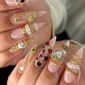 May include: Close-up of long, pointed fingernails with a light pink base. The nails are decorated with gold chains, pearls, and red and clear gemstones. Some nails feature miniature watch faces, creating a luxurious, ornate design.