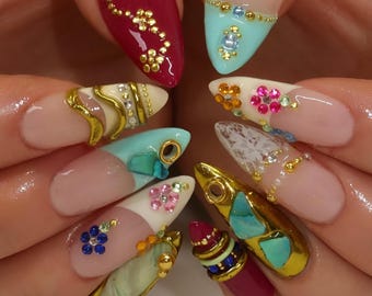 May include: Almond-shaped fingernails featuring a diverse array of nail art designs. Decorations include glossy red and mint green polish, gold metallic accents, small colorful rhinestones, white lace patterns, and embedded turquoise-colored stones.