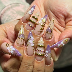 May include: A hand showing long, almond-shaped artificial nails. The nails are painted with a nude base and elaborate designs. Gold accents, purple tips, and white floral decorations adorn the nails, creating a glamorous and artistic appearance.