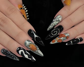 May include: Long stiletto-shaped nails featuring a mix of black, white, silver, and orange accents. Designs include metallic silver textures, raised orange blobs, black and white swirls, and silver spikes on translucent orange nail tips.