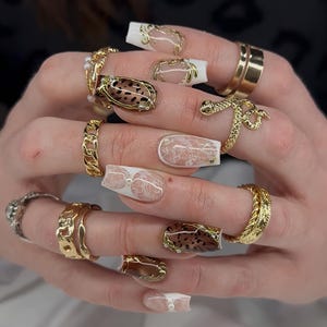 Puede incluir: Primer plano de manos adornadas con anillos de oro y uñas largas y decoradas. Los anillos presentan varios diseños, incluyendo una serpiente, eslabones de cadena y bandas texturizadas. Las uñas están pintadas con patrones blancos, dorados y marrones, incluyendo estampado de leopardo y encaje.