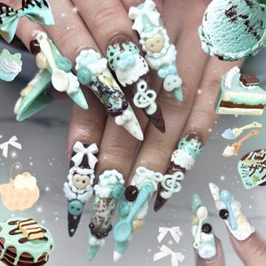 Puede incluir: Uñas largas de estilo estilete con diseños 3D de conos de helado en verde menta y marrón, lazos blancos, cucharas pequeñas y notas musicales. El arte de uñas incluye texturas de chocolate, dijes de corazón y botones, creando una estética de postre en capas con varios gráficos de dulces alrededor de los dedos.