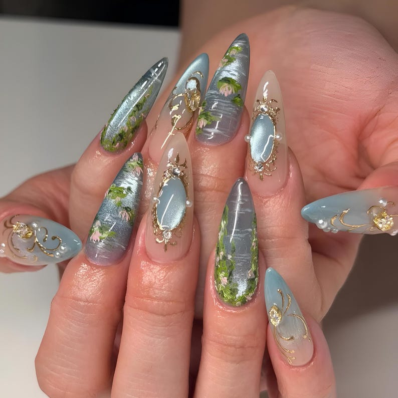 May include: Long, almond-shaped artificial nails with a light blue, silver, and clear design. Some nails feature a water scene with green and pink accents, while others have gold filigree and pearl embellishments.