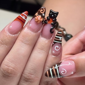 May include: Almond-shaped artificial nails featuring a nude base with brown, orange, and white tips. Designs include horizontal stripes, white swirls, a small 3D orange pumpkin, a black cat figurine, and tiny white pearl accents on each nail.