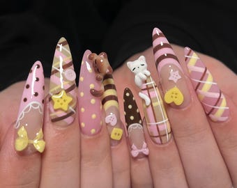 May include: A set of long, pointed acrylic nails with various designs. The nails feature pink, brown, and yellow patterns, including polka dots, plaid, and stripes. Some nails have 3D embellishments like a teddy bear, a cat, buttons, stars, and bows.