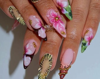May include: Stiletto-shaped nails featuring a mix of designs including hand-painted pink floral motifs, green leaf patterns, and black tiger-like stripes. The nail art is accented with small gold metallic beads and clear rhinestones.